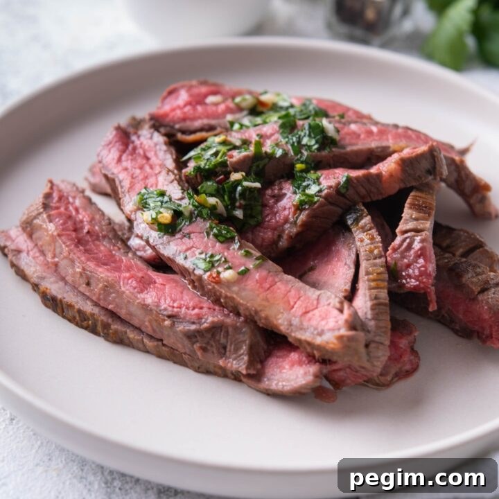 Thinly sliced bavette steak piled on a white plate and covered in chimichurri sauce.