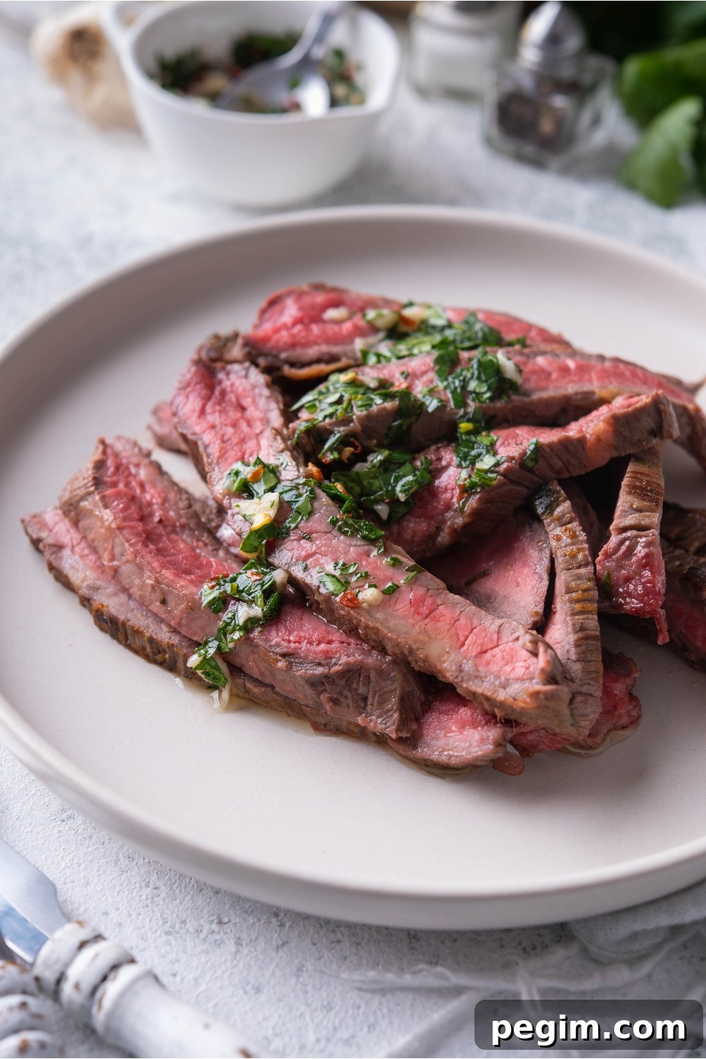 Thinly sliced bavette steak piled on a white plate and covered in chimichurri sauce.