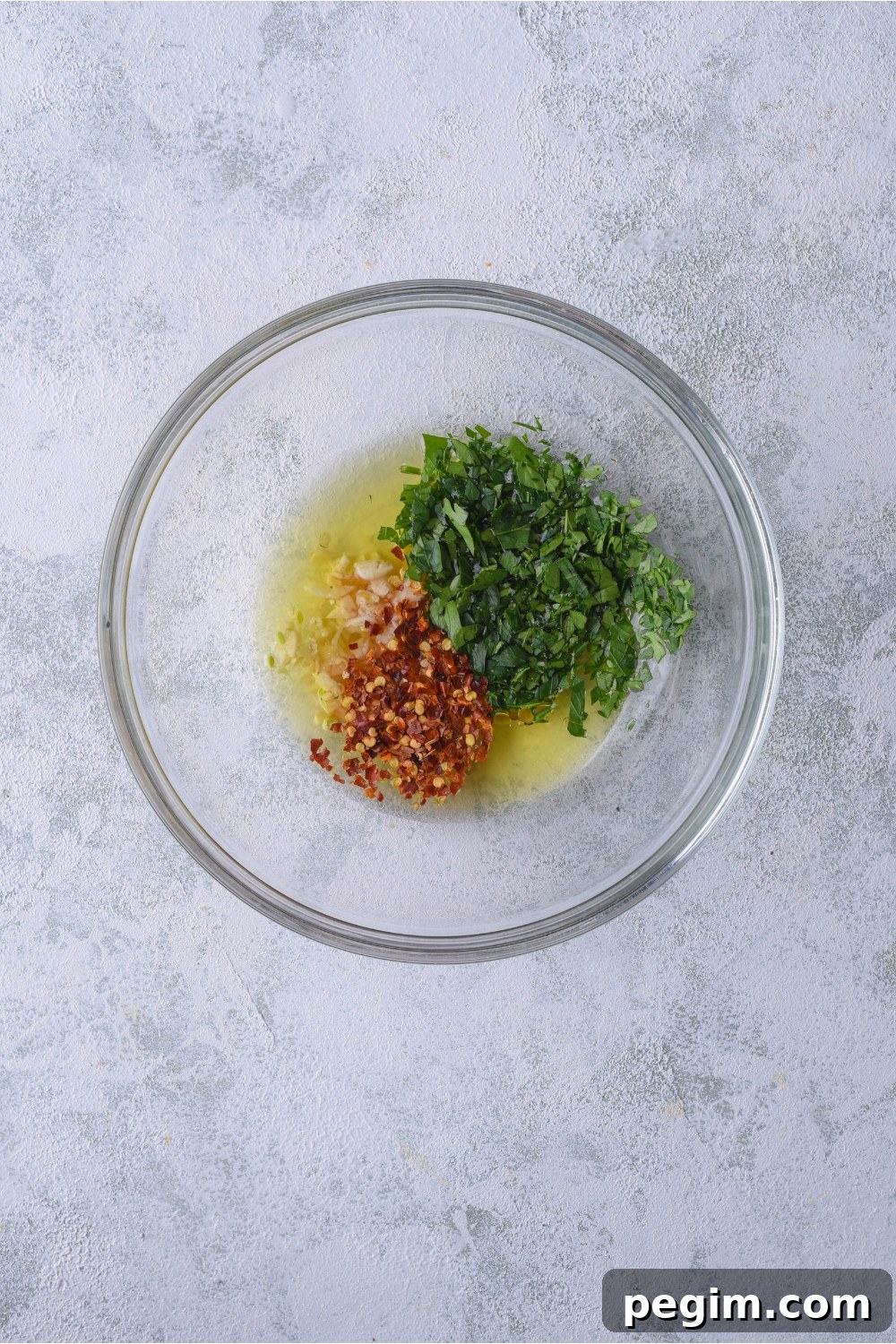 A clear bowl filled with oil, garlic, chopped green herbs, and red chili flakes.
