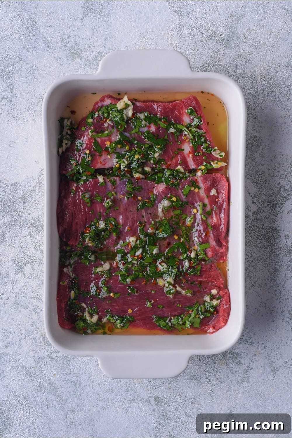 A white baking dish filled with bavette steaks marinating in a blend of green herbs.