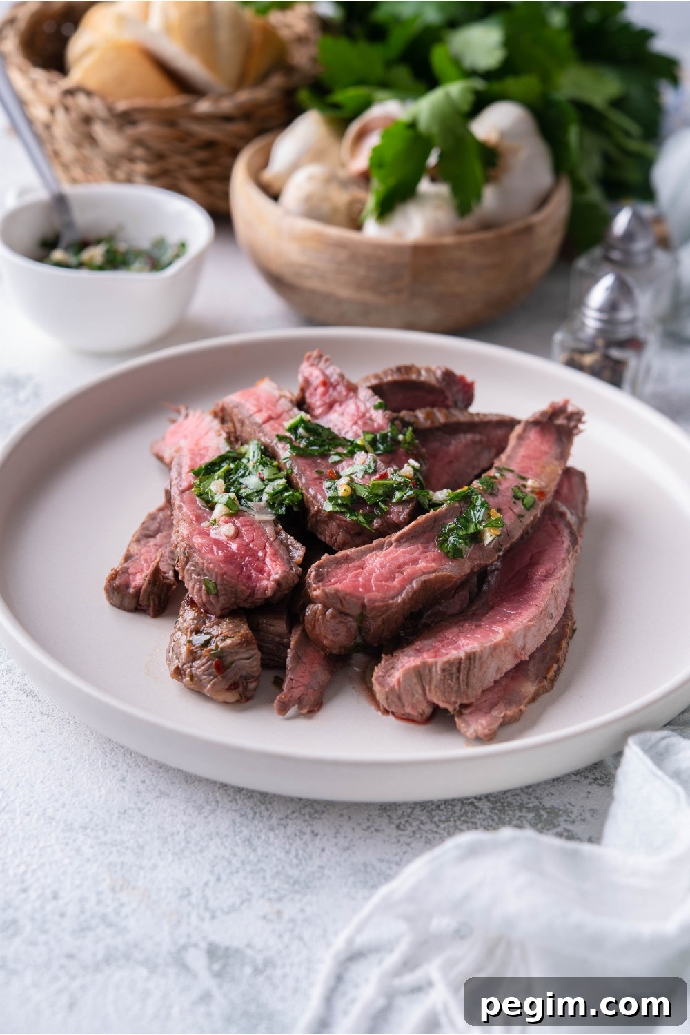 Thinly sliced bavette steak piled on a white plate and covered in chimichurri sauce.