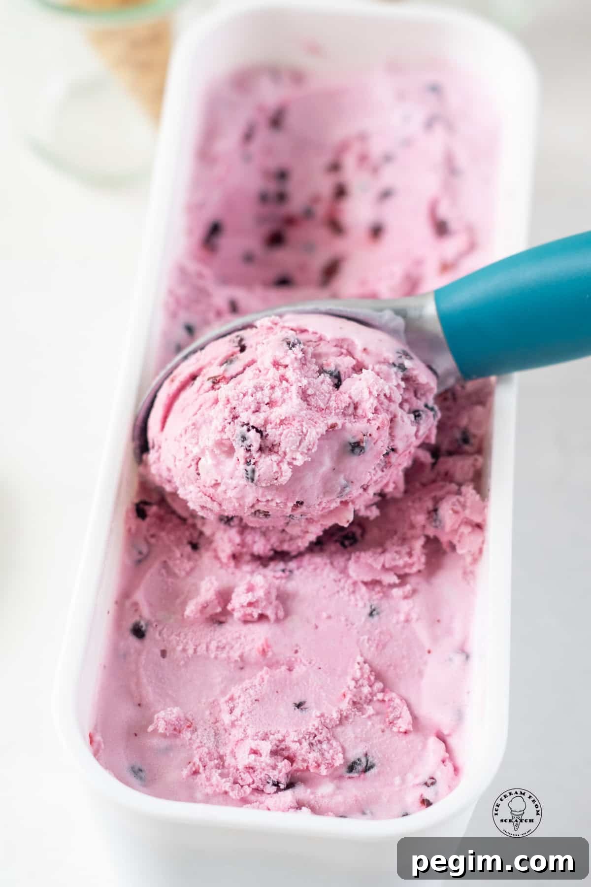 a blue ice cream scoop scooping huckleberry ice cream from a white plastic container.