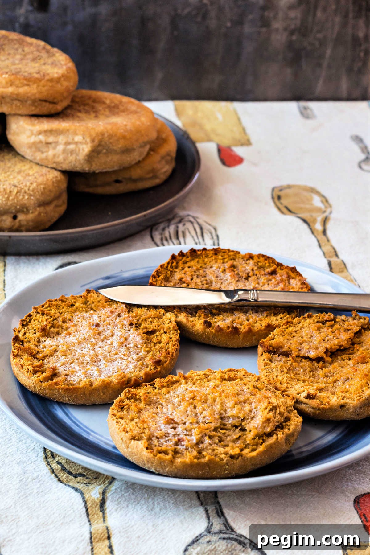Two split, toasted, and buttered English muffins on a plate with another plate of unsplit muffins in the background.