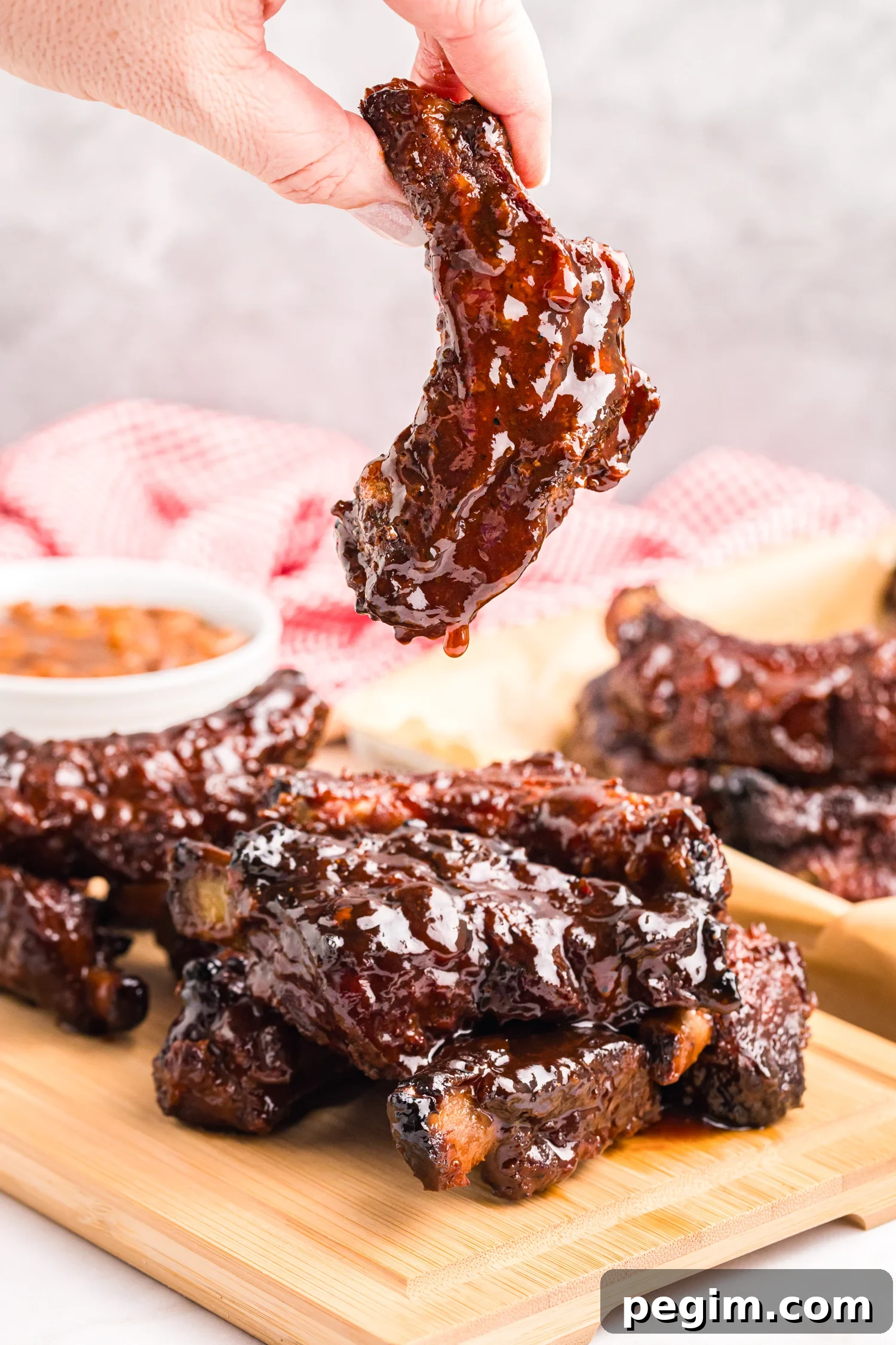 A hand holding a single barbecue-glazed rib dripping sauce above a stack of ribs on a wooden board.
