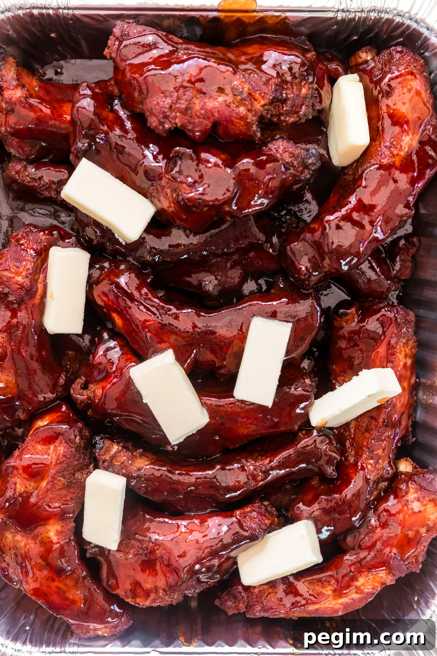 Smoked pork ribs coated in glossy barbecue sauce with slices of butter, resting in an aluminum pan.