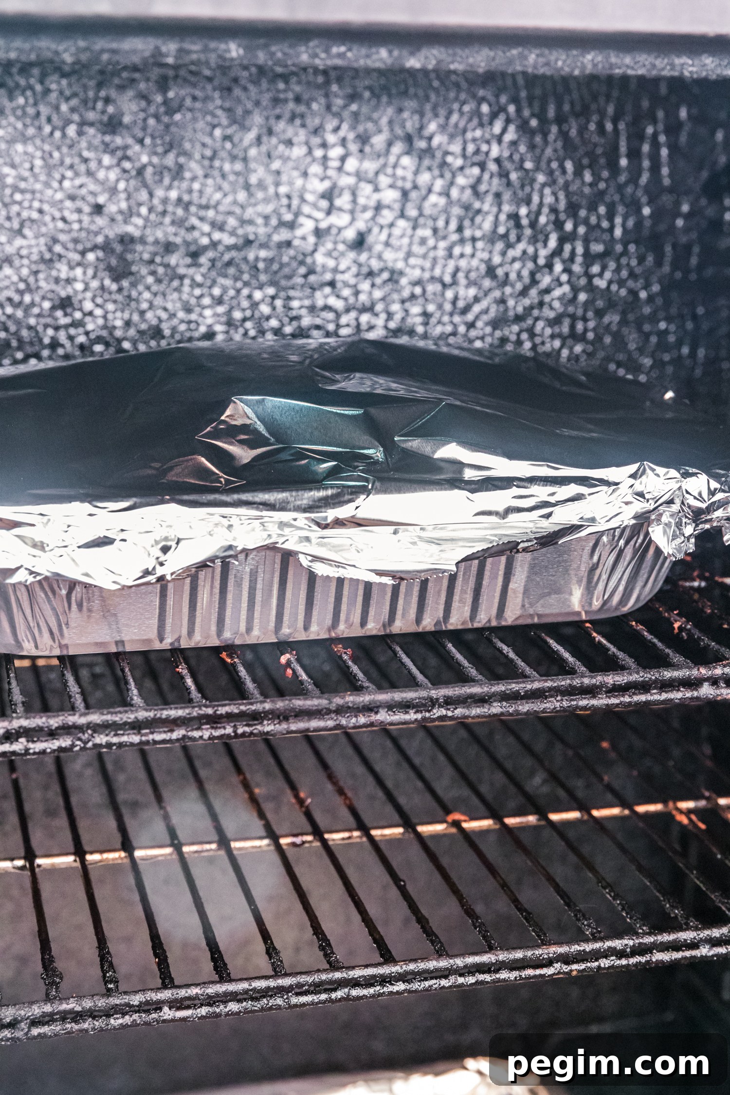 Foil-covered aluminum pan of ribs placed inside a smoker on metal grates.