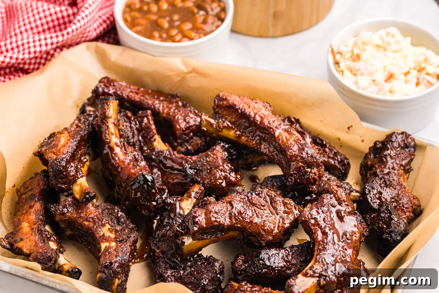 Pile of sticky barbecue pork ribs served on parchment paper with baked beans and coleslaw in the background.
