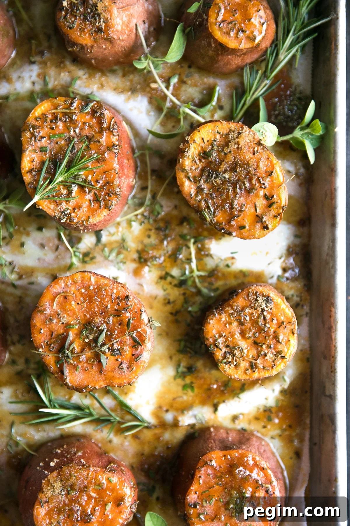 Roasted sweet potatoes on a large baking sheet covered with melted butter, brown sugar, and fresh herbs.