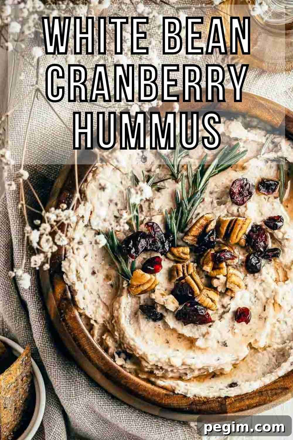 Overhead shot of hummus garnished with pecans, cranberries, and rosemary in a large wooden bowl on a burlap cloth with rosemary, baby breath, and crackers on the side.