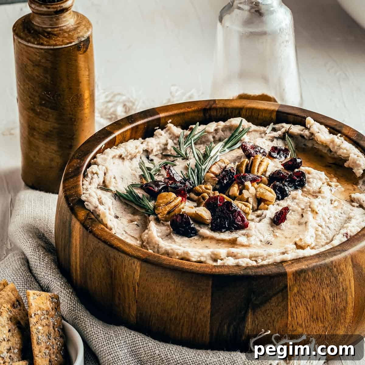 Side shot of hummus garnished with rosemary, pecans, and cranberries in a large wooden bowl on a burlap cloth with crackers, and two jars in the background.