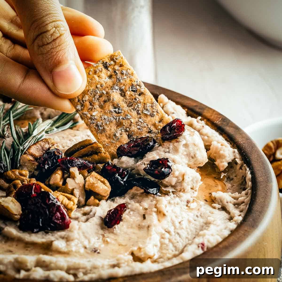 Side close up of a hand scooping some hummus with a cracker - the hummus is garnished with rosemary, cranberries, and pecans.
