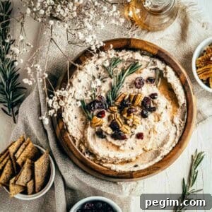Overhead shot of hummus garnished with pecans, cranberries, and rosemary in a large wooden bowl on a burlap cloth with rosemary, baby breath, and crackers on the side.