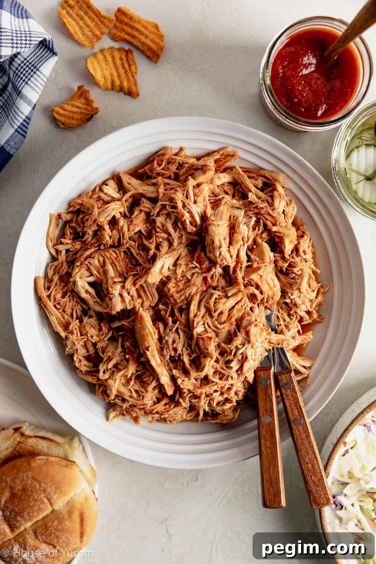 Shredded BBQ Chicken in a bowl with hamburger buns, pickles and sauce next to it.