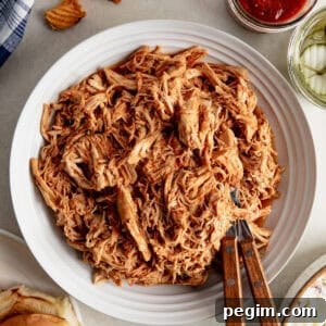Shredded BBQ Chicken in a bowl with hamburger buns, pickles and sauce next to it.