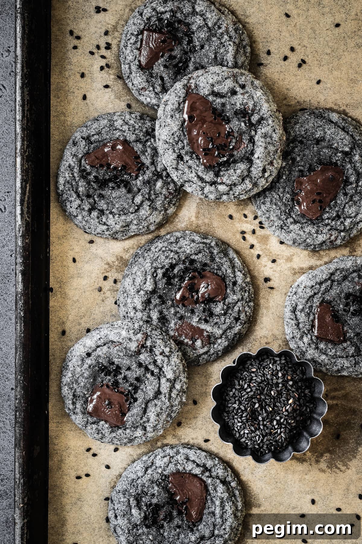 Grey cookies with pools of melted chocolate and sesame seeds on a parchment lined baking sheet.