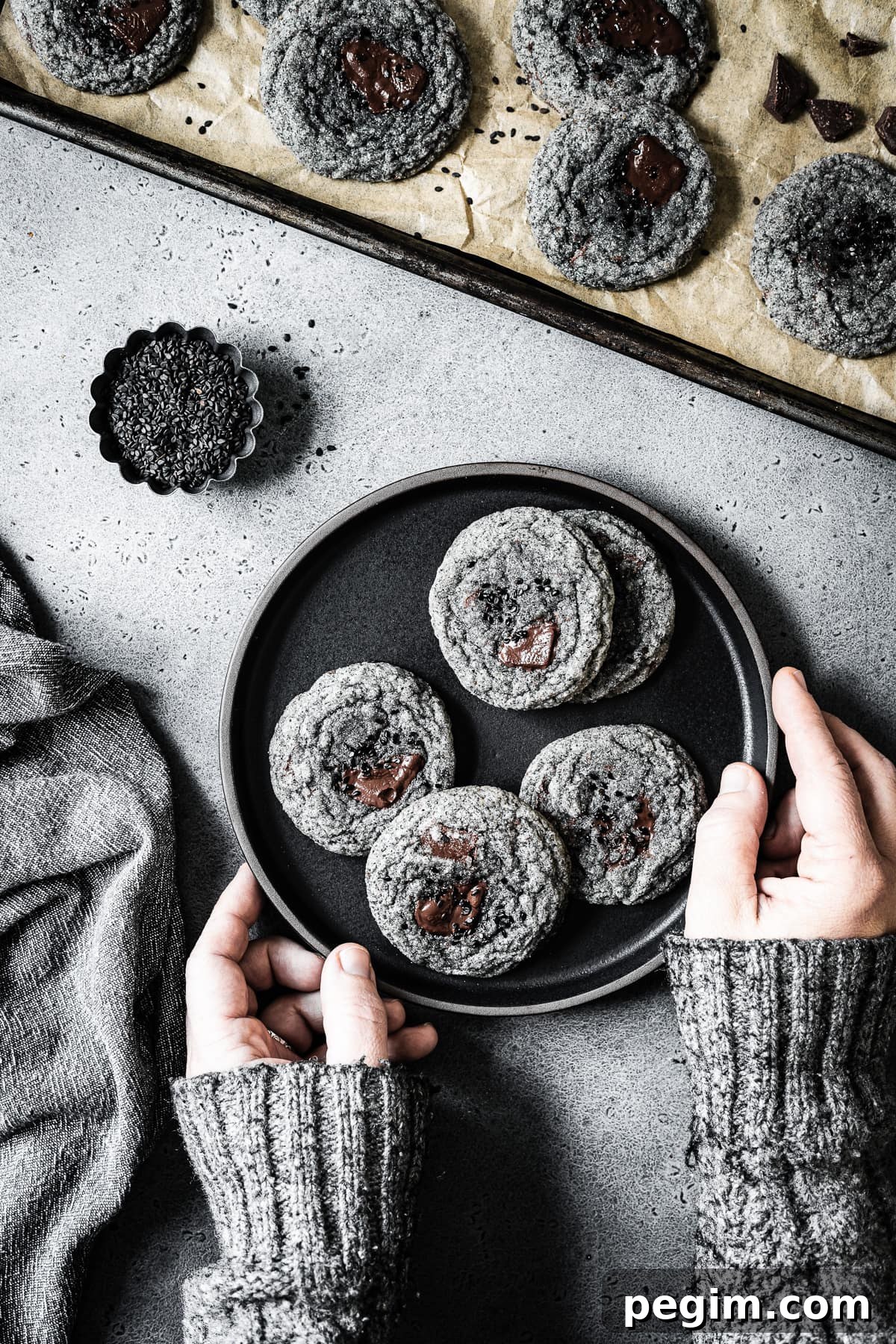 Arms in a grey sweater reach into the frame to hold a black plate of cookies. The colors in the image are monochromatic: grey, black and brown. The sweater color and texture matches the grey stone background and the color of the cookies.