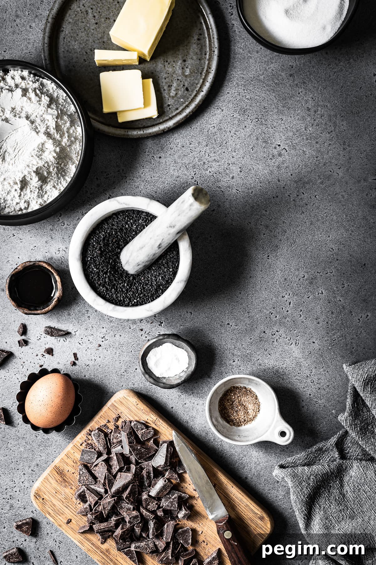 Various bowls and containers filled with ingredients for making cookies arranged on a grey stone background. The light is entering from left. The image is well lit and there are dark shadows.