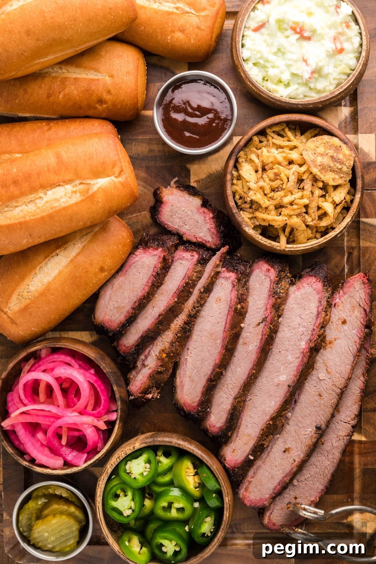 Sliced brisket, hoagie rolls, coleslaw, pickled red onions, jalapeños and barbecue sauce on a board.