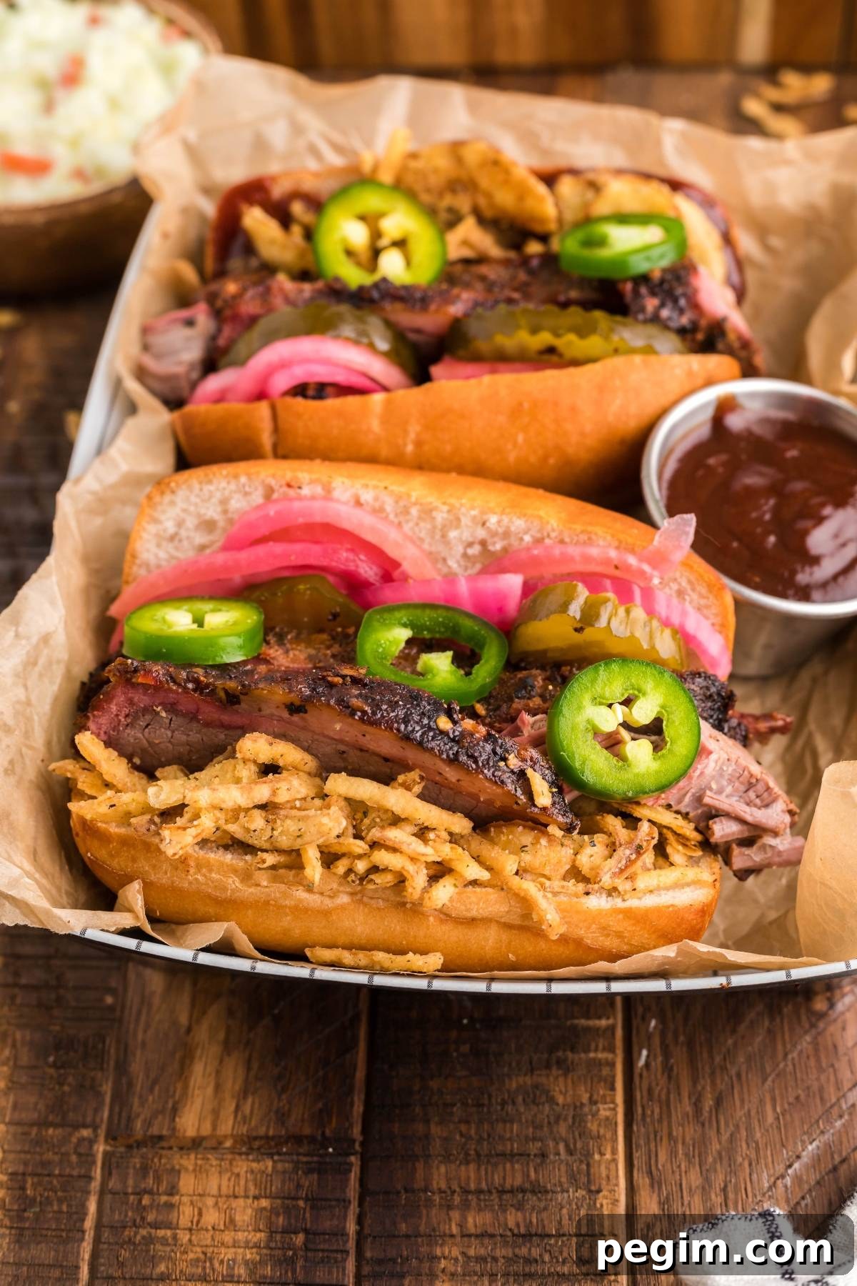 Two BBQ brisket sandwiches with pickled red onions, jalapeños and crispy onions on a tray with BBQ sauce.