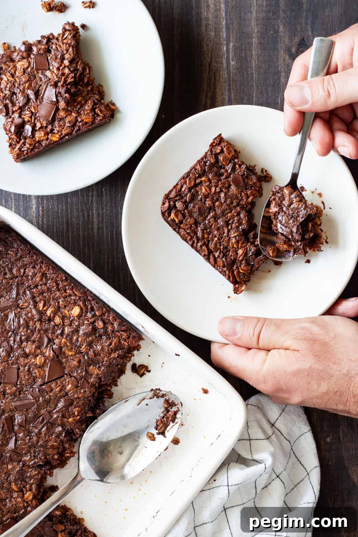 Chocolate Baked Oatmeal in a baking dish with two portions on small plates and a hand taking a spoonful from one portion.
