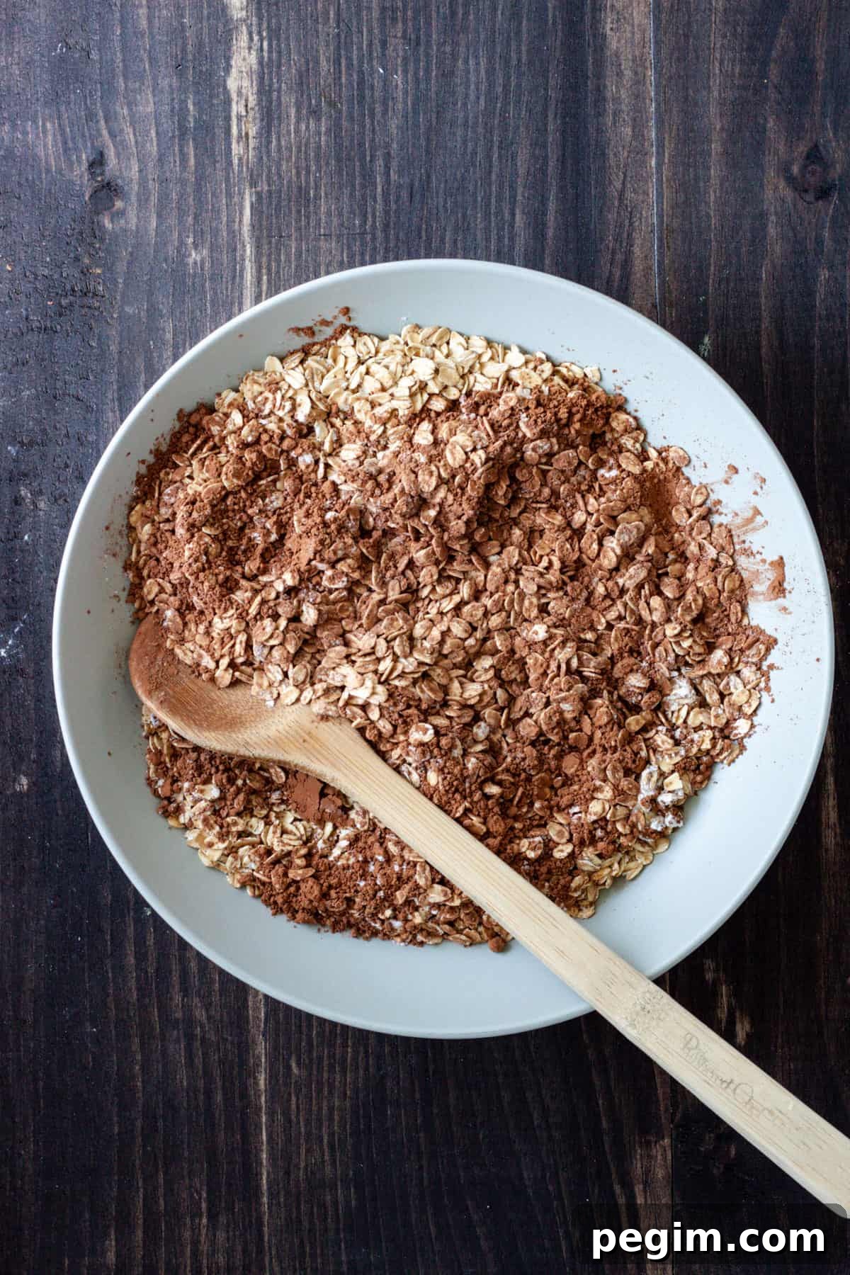 Oats and cocoa powder in a mixing bowl with a wooden spoon getting ready to mix it all together.