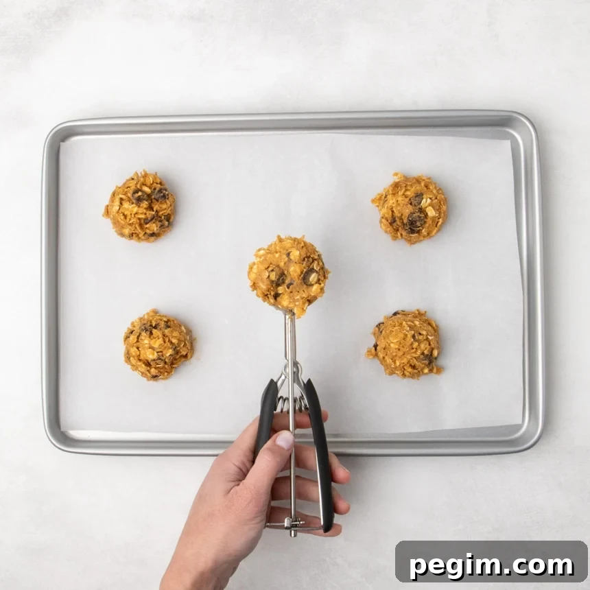 A hand holding a black-handled cookie scoop with raw cookie dough above a parchment-lined baking sheet with four other portions of raw rust-colored oatmeal cookie dough spaced apart.