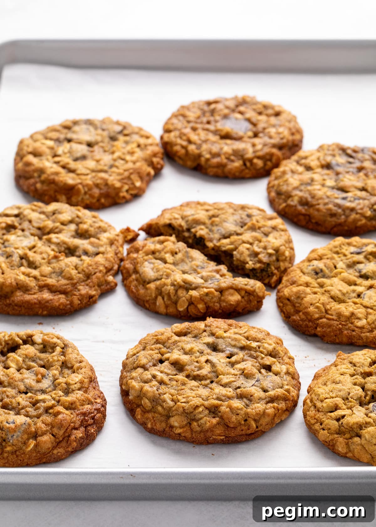 Nine oatmeal cookies on a parchment-lined metal baking sheet, with one cookie broken in half near the center.