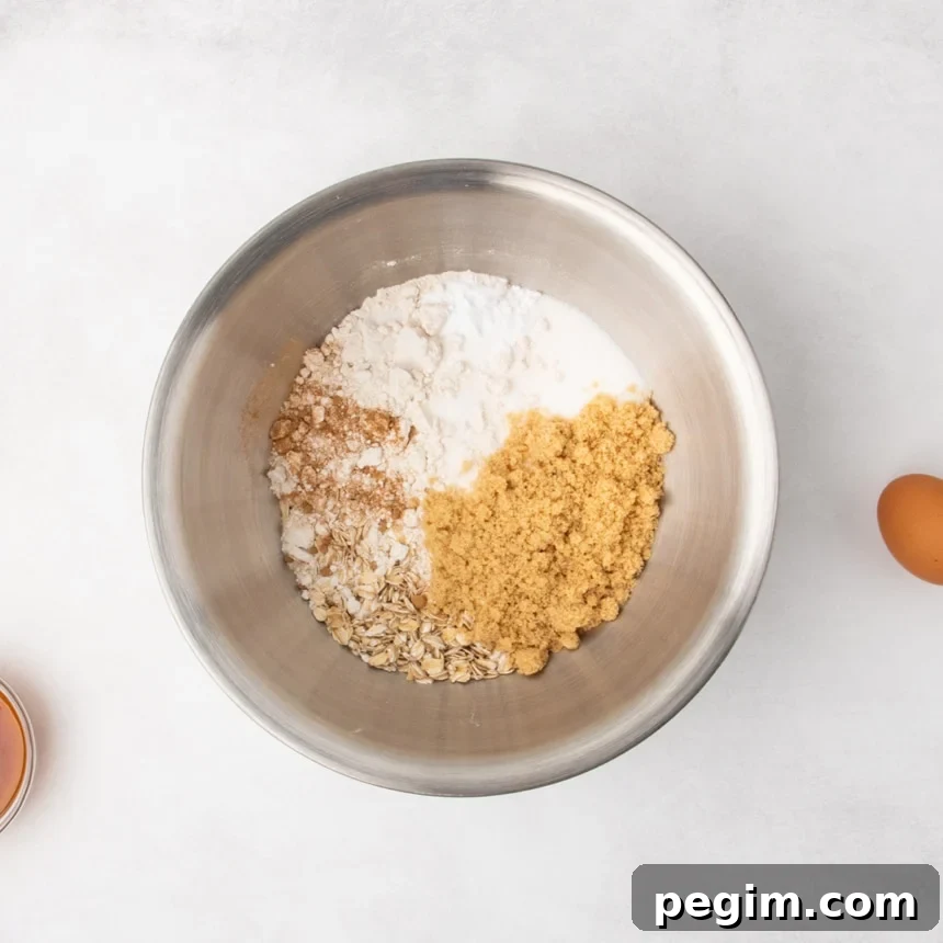 A metal mixing bowl containing unmixed rolled oats, brown sugar, white sugar, and baking soda and light brown powder on a light gray surface, with an egg and a small bowl partially visible nearby.