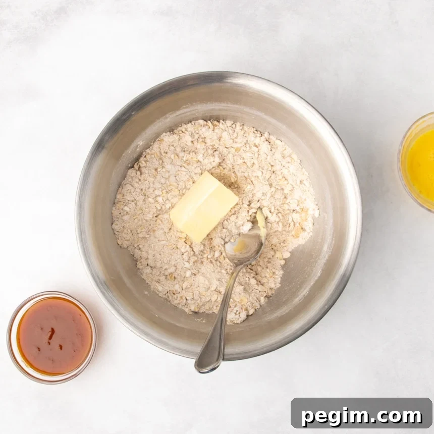 A metal mixing bowl filled with a mixture of pale brown dry ingredients and a piece of butter on top, with a metal mixing spoon resting inside the bowl; two small bowls, one containing orange puree and one with yellow beaten egg, are nearby.