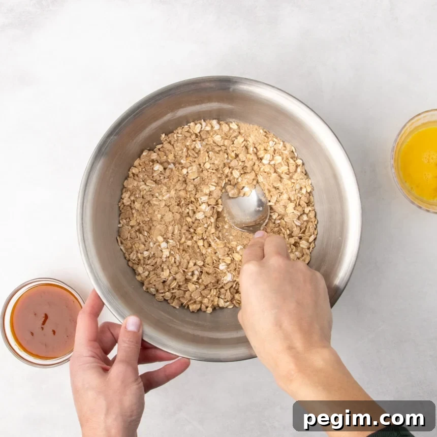 A person’s hands holding a metal spoon while mixing dry ingredients with oats in a metal bowl; small bowls of orange puree and a yellow beaten egg are nearby.