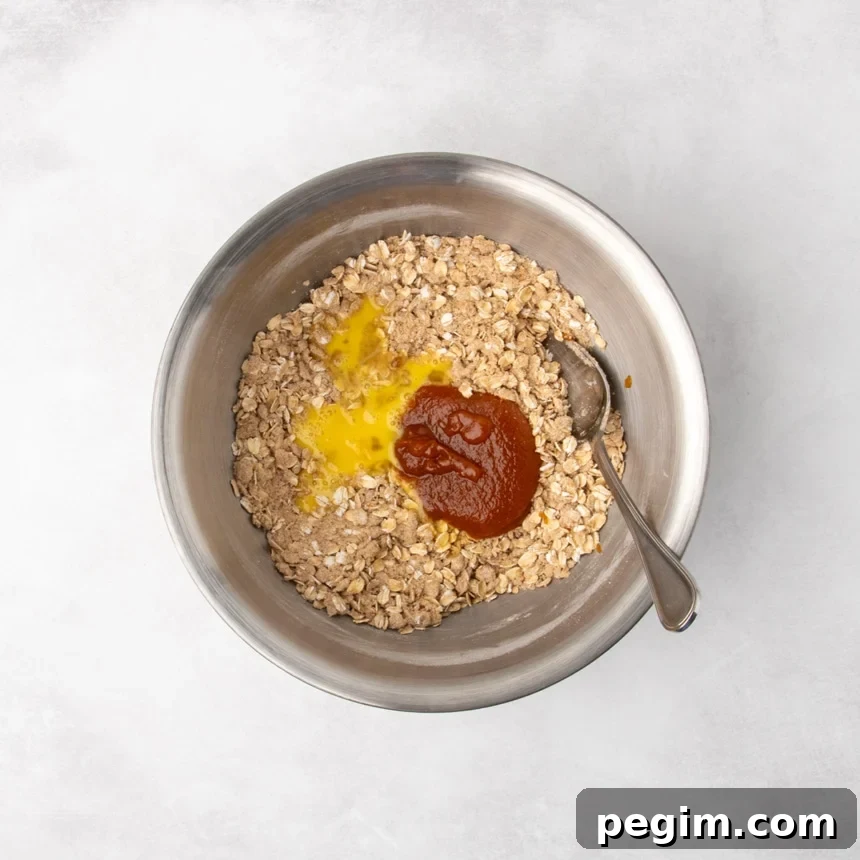 A metal mixing bowl containing a mixture of oats and dry ingredients with a beaten egg and orange pumpkin butter puree added on top, unmixed.