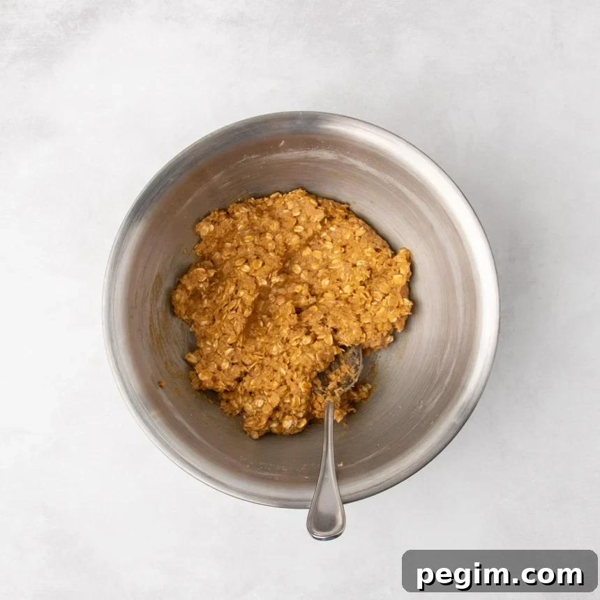 A metal mixing bowl containing combined rusty brown colored raw oatmeal cookie dough with a spoon resting inside.
