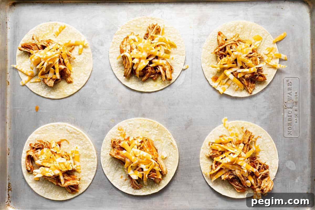 Small corn tortillas topped with shredded chicken and shredded cheese on a metal baking sheet.
