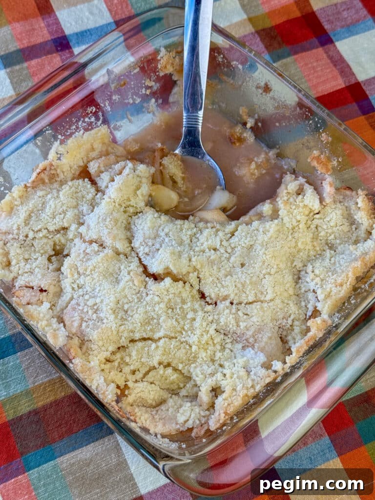 Overhead view of a freshly baked old fashioned apple crisp recipe with a spoon ready to scoop out another serving out on an orange and beige plaid napkin.