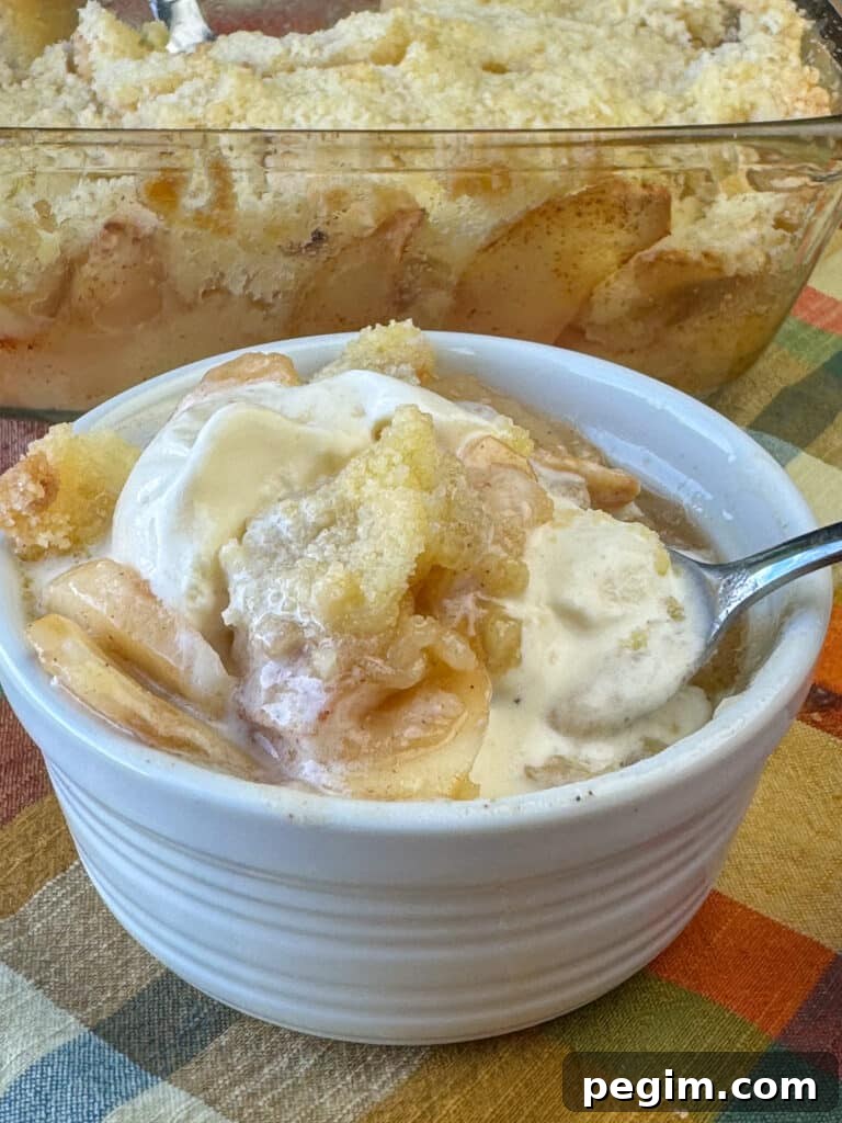 Spoon lifting delicious slices of apples in an easy apple crisp with melting ice cream out of a small white dish with the full baking dish in the background.