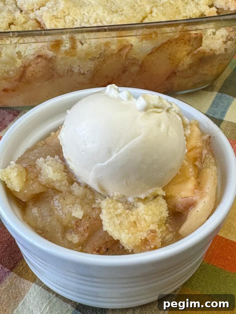 Close up of a crunchy baked topping on a small bowl of old fashioned apple crisp garnished with ice cream and the full baking pan in the background.