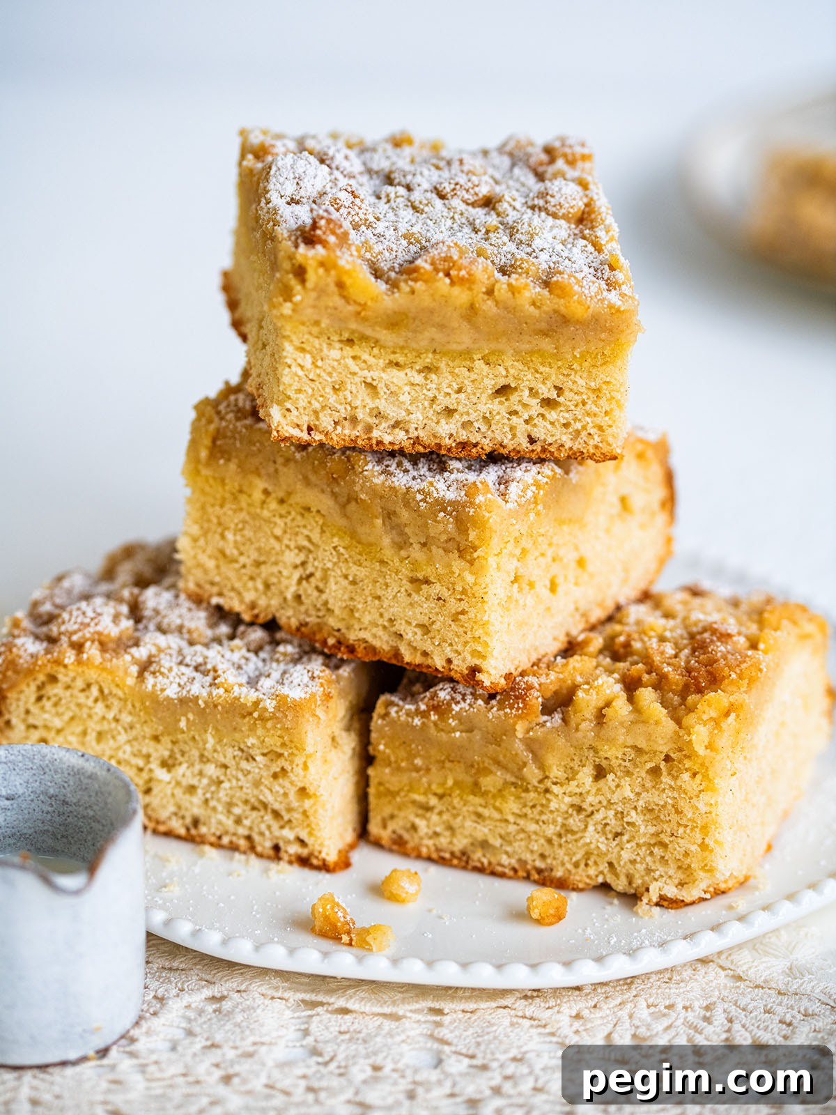 Authentic German Streuselkuchen Recipe: Classic Crumb Cake Guide 3 Four square pieces of German crumb cake (streuselkuchen) stacked on a white plate, dusted with powdered sugar. The cakes feature a golden brown crumb layer and a soft, fluffy base. A small pitcher rests nearby on a textured tablecloth.