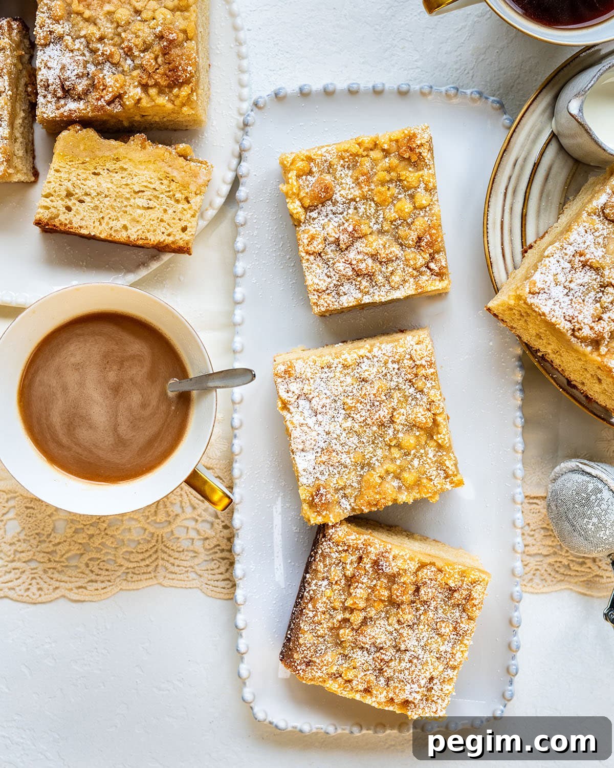 Authentic German Streuselkuchen Recipe: Classic Crumb Cake Guide 5 Three slices of crumb cake topped with powdered sugar on a white tray, next to a cup of coffee, a small pitcher of cream, and additional cake slices on a plate, all on a light table setting.