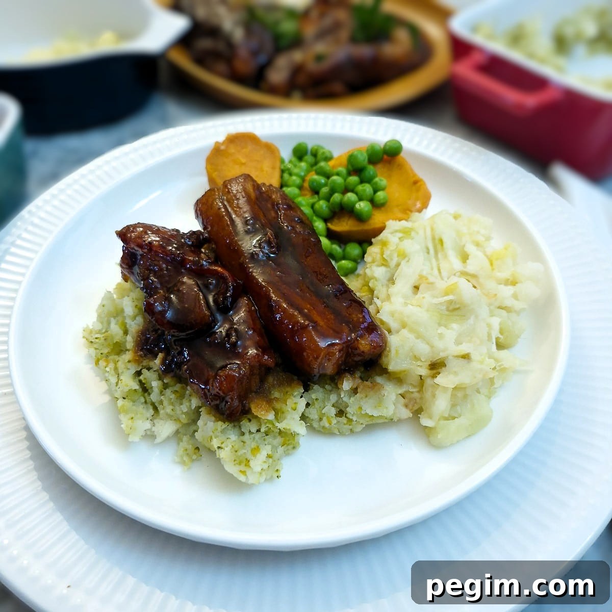 Two glazed pork belly slices on a pile of vegetable mash on a white plate, with sweet potatoes and peas.