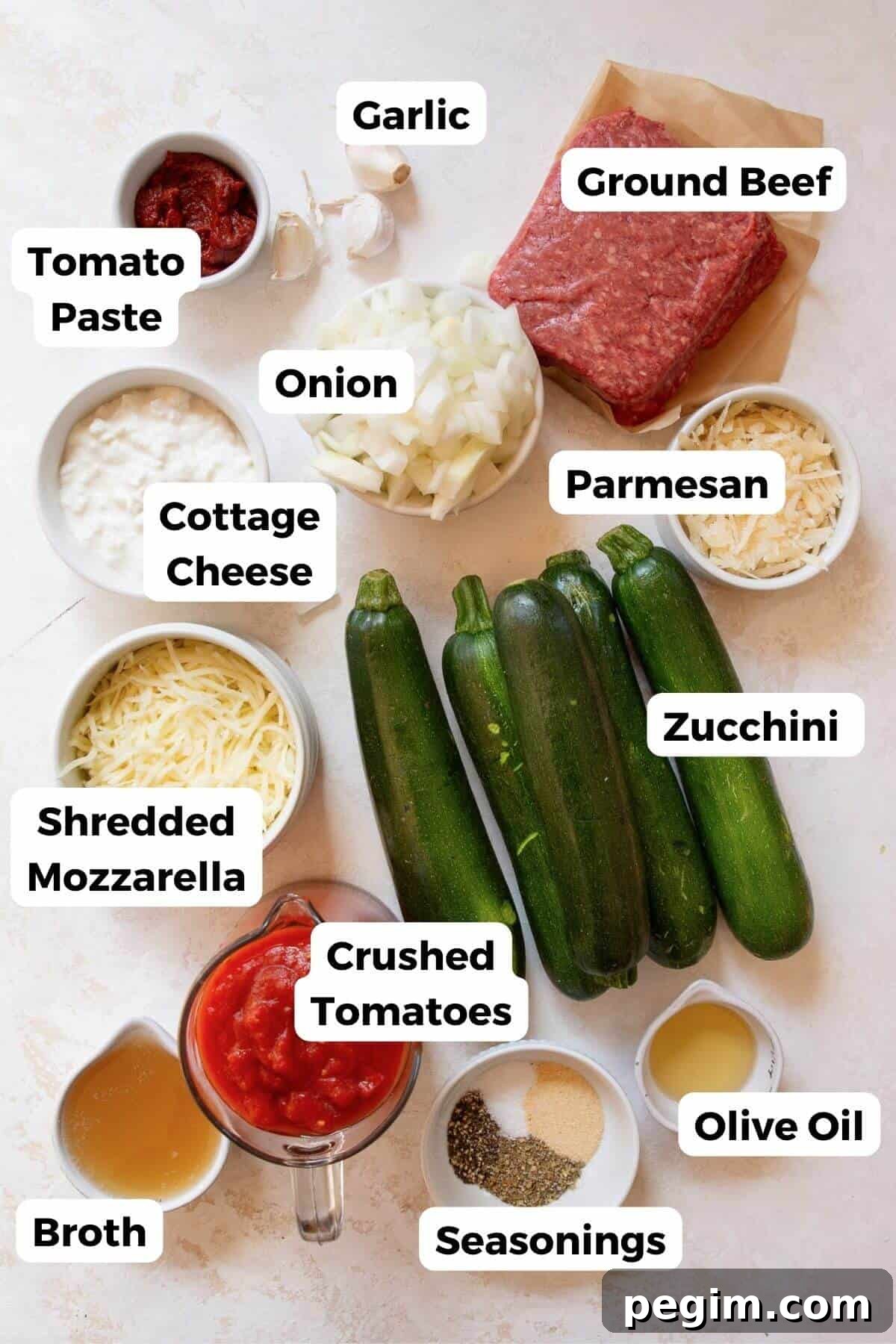 All ingredients in ground beef and zucchini casserole in small bowls with labels - zucchini, lean ground beef, onion, garlic, crushed tomatoes, tomato paste, cottage cheese, parmesan, mozzarella, avocado oil, Italian seasoning, garlic powder, salt, pepper, broth, and fresh basil.