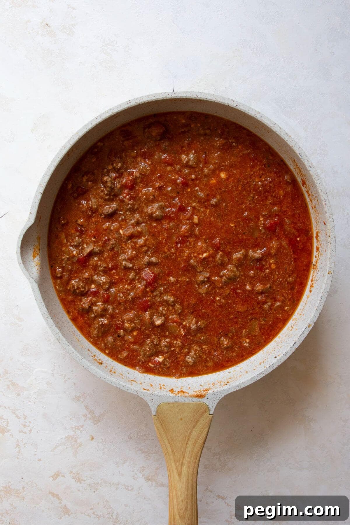 Adding crushed tomatoes, cottage cheese, parmesan, tomato paste, and broth to cooked ground beef in a skillet.