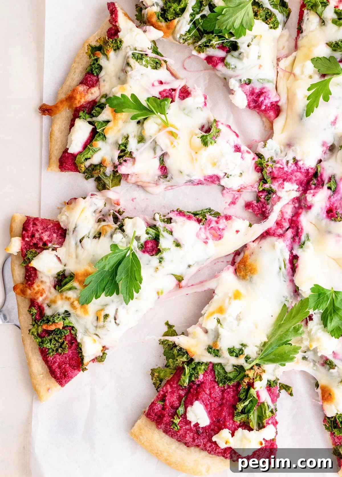 Pizza cut into slices on a sheet of parchment paper with beet pesto sauce, kale, and goat cheese.