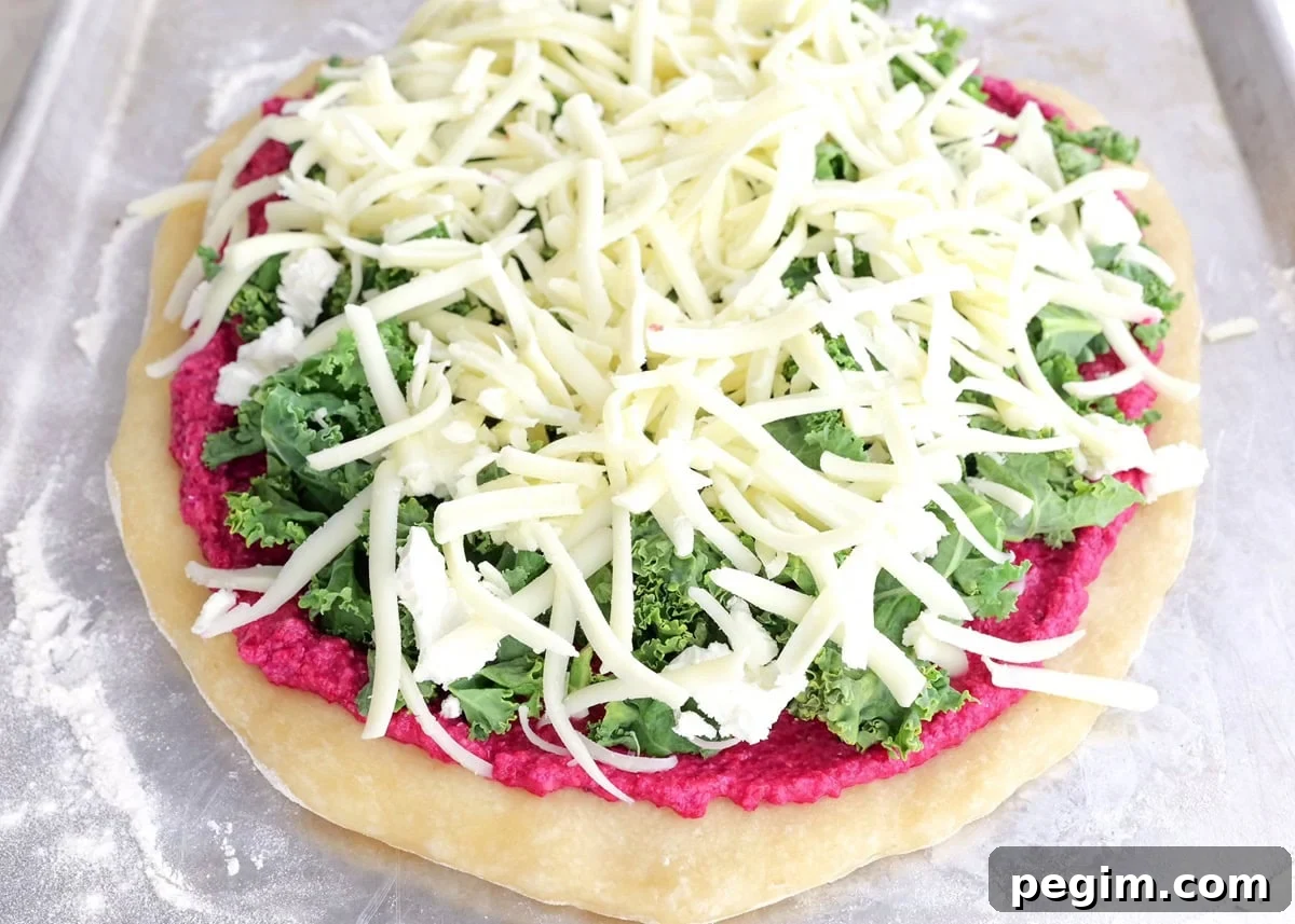 Pizza crust with beet pesto sauce, chopped kale, goat cheese, and mozzarella cheese on top, ready to go into the oven.