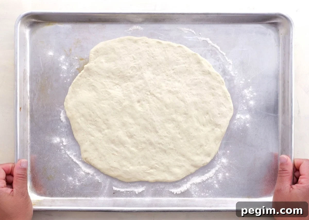 Pizza dough spread out onto a baking sheet with flour beneath it, ready to go into the oven to prebake.