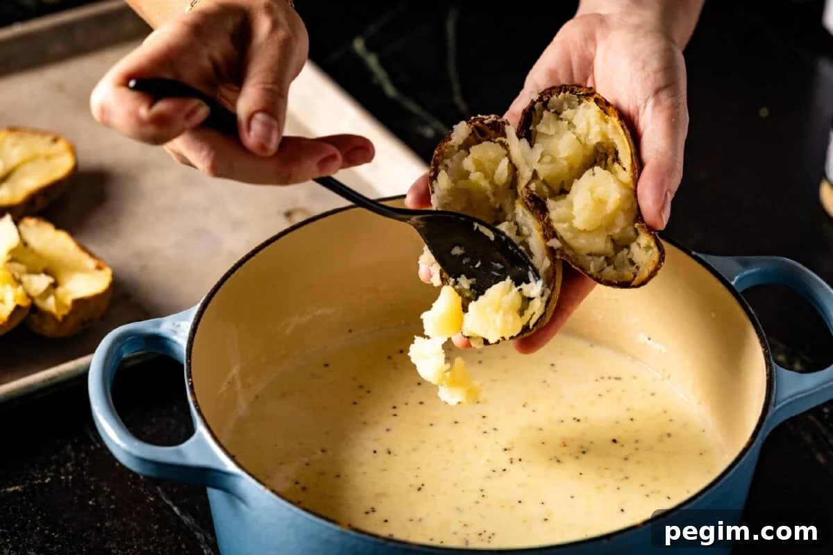 A chef's hand scooping the inside of a potato into a large pot of liquid using a spoon.