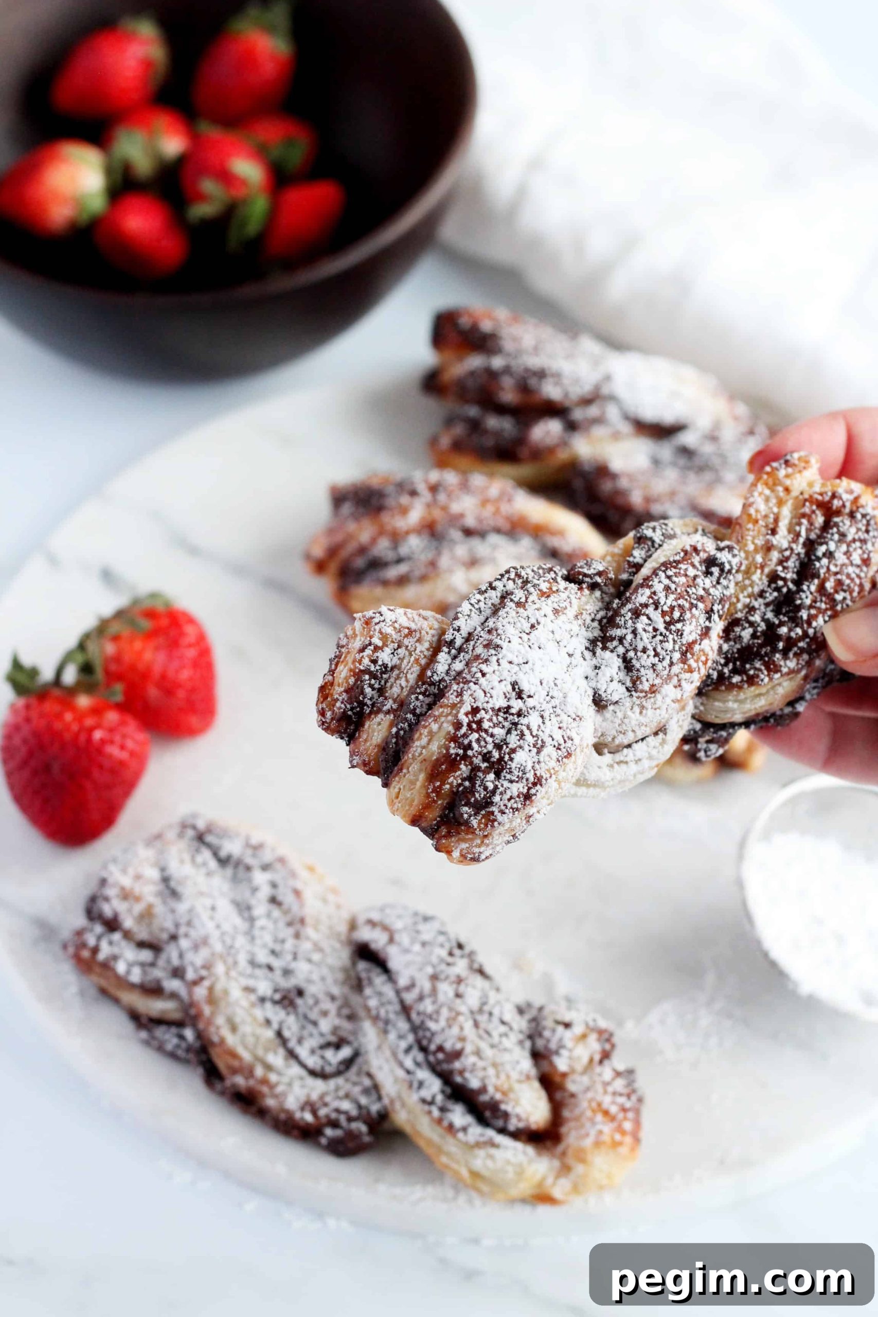 Nutella puff pastry twists dusted with powdered sugar, on marble plate with strawberries in the background