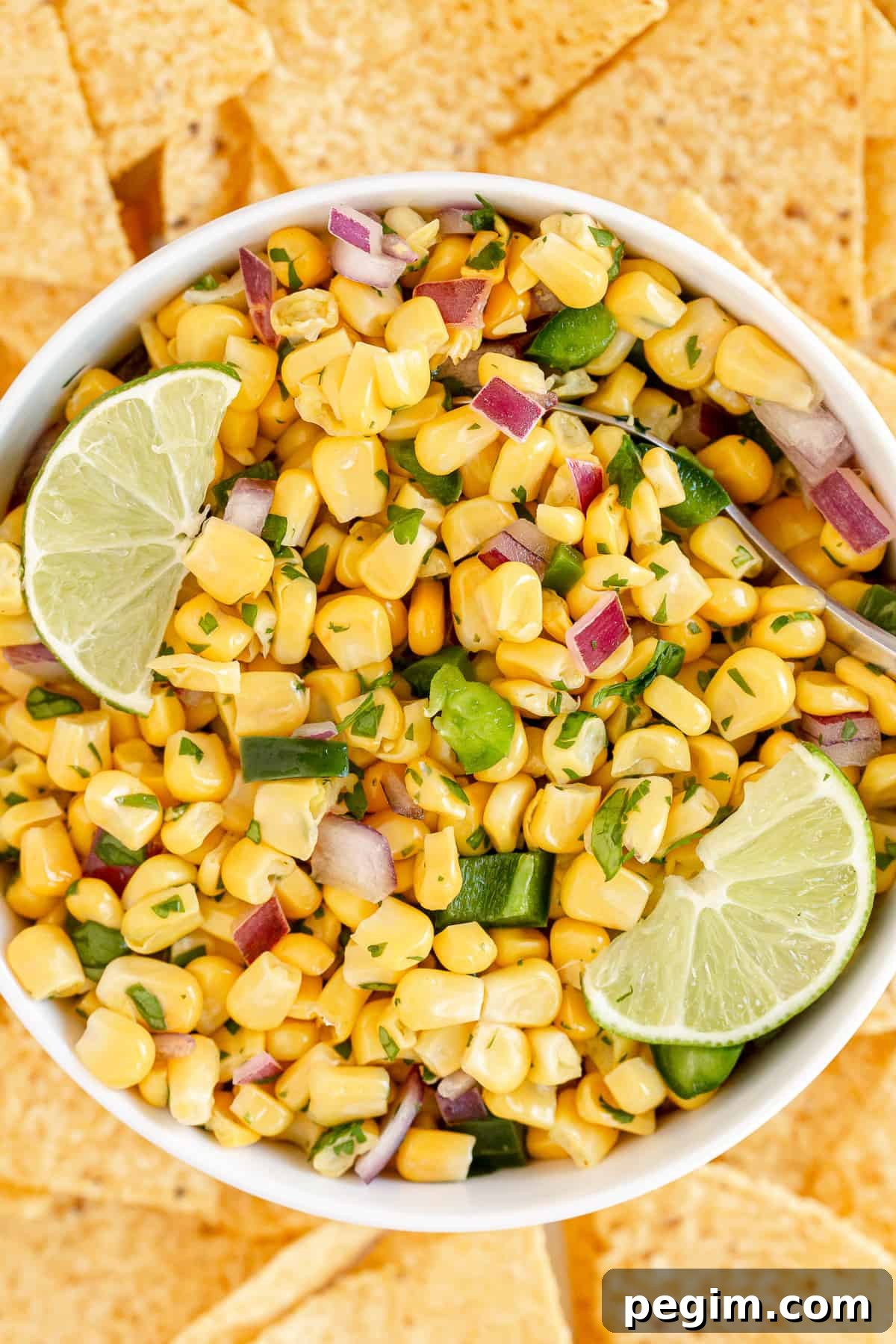 Corn salsa in a white bowl with lime wedges surrounded by tortilla chips.