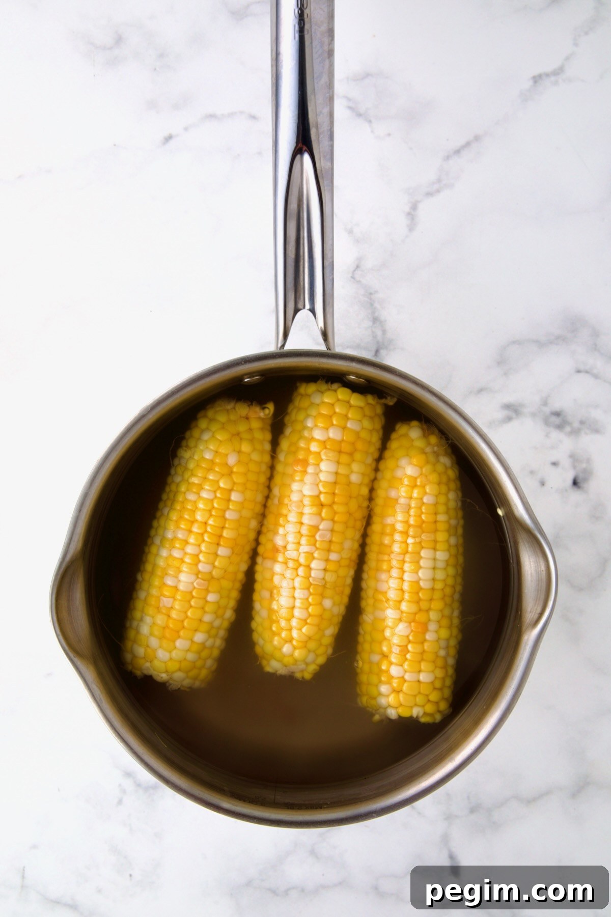 Three ears of corn in boiling water in stainless steel saucepan.