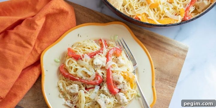 Strips of onion and red and yellow bell peppers with diced chicken on a bed of angel hair pasta on a white plate with a yellow rim. The plate is on a wooden cutton board on a white marble counter. There is an orange napkin beside the plate. At the top of the photo is part of a skillet with angel hair pasta, strips of red and yellow bell peppers, and diced chicken.