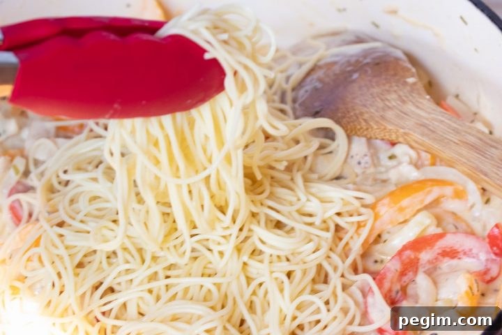 A wooden spoon stirring sliced onions and red and yellow bell peppers and diced chicken, tarragon, half and half, and shredded mozzarella cheese in a skillet. Cooked angel hair pasta is on top of the vegtables and cheese and a pair of red tongs is holding some of the pasta above the skillet.